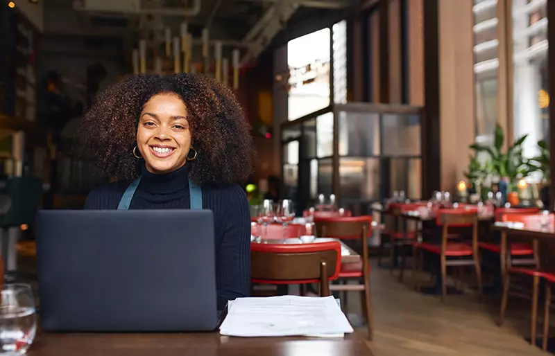 一位系着围裙的业务经理坐在餐厅的餐桌前，使用着一台笔记本电脑