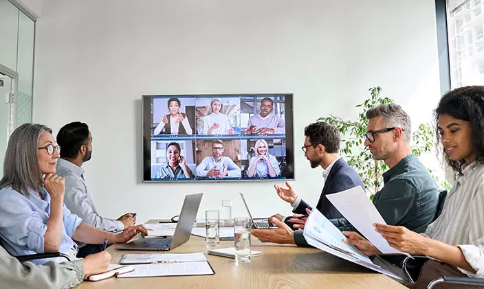 Group of diverse business professionals at a table speaking with other employees on a monitor hanging on the wall.