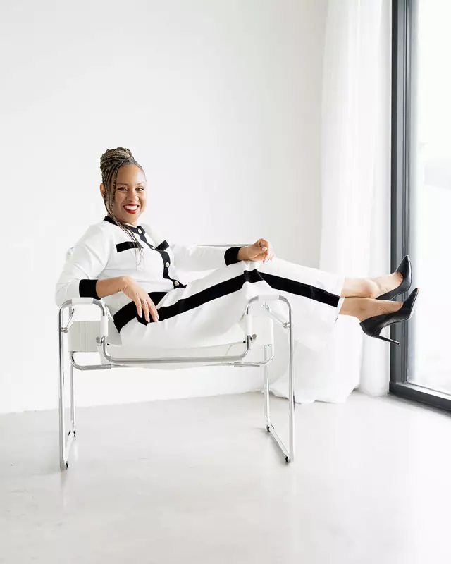 Freda Richards smiling while seated in a modern chair with her legs up, wearing a white and black outfit. She exudes confidence and calm.