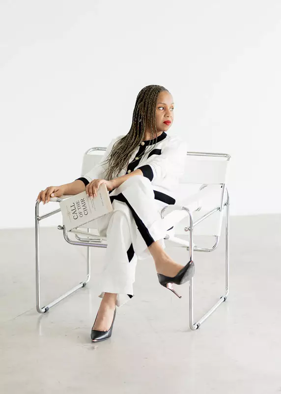 Freda Richards sitting in a modern chair, holding a book titled “Call of the Chair,” looking thoughtful.