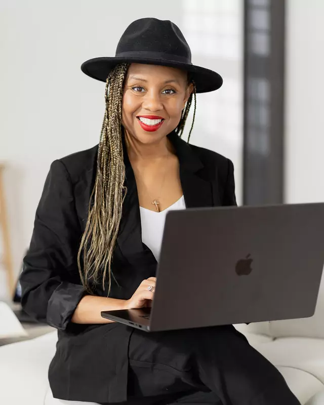 Freda Richards wearing a black hat and blazer, smiling as she works on a laptop in a bright studio.