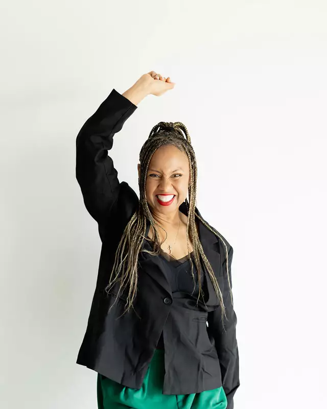 Freda Richards raising a fist and smiling in a confident pose, wearing a black blazer and green pants.