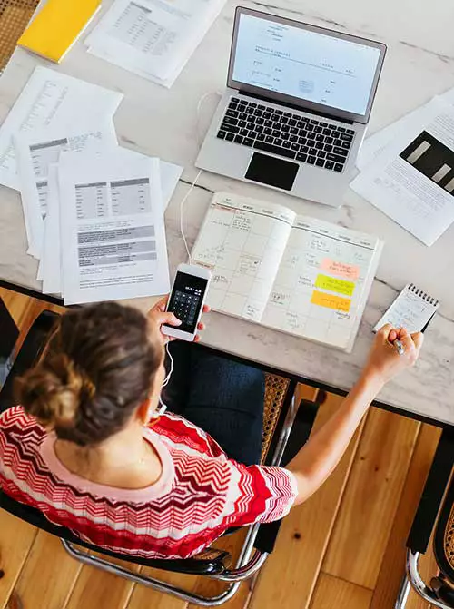 An accountant, viewed from overhead, seated at a desk covered with papers and a laptop