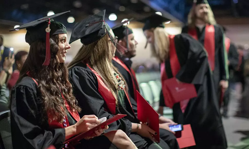 University of Phoenix students looking eager in cap and gown getting seated for a graduation ceremony.