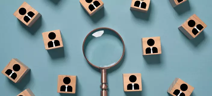 A magnifying glass surrounded by blocks with people icons on them to signify using a skills gap analysis.