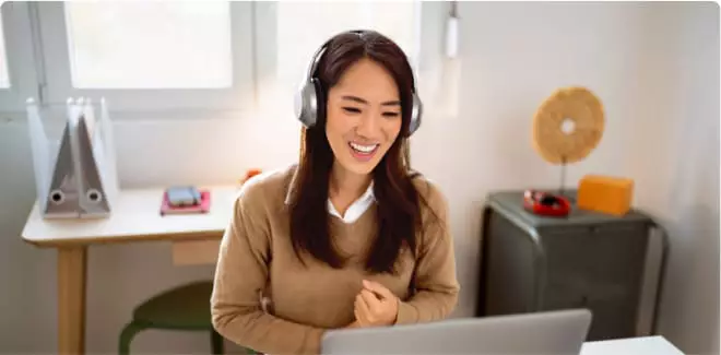 A woman wearing headphones is seated at a laptop, smiling and talking with colleagues online