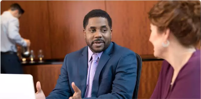 A man dressed in a suit and tie chats with a colleague in a conference room