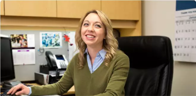 A woman sits a cubicle, smiling and talking with a colleague standing at her desk