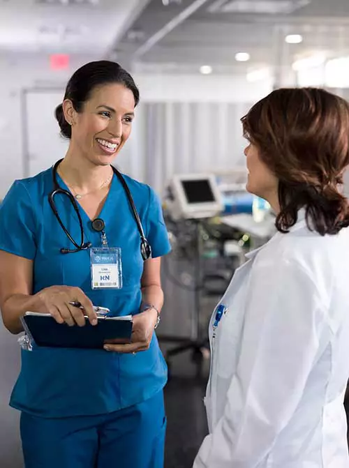 Two medical professionals smile and talk in a hospital hallway