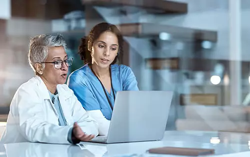 Two medical professionals look at a laptop screen and discuss a patient's chart