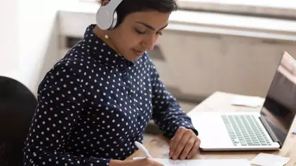 Student with headphones at a desk takes notes on a sheet of paper