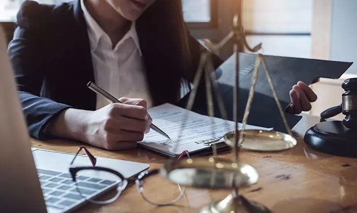 Photo of a female professional working hard on a legal document in front of 'justice' scales. Top of face is cropped out.