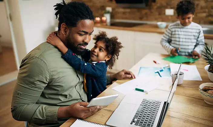 Father taking online classes at home while young child hugs him