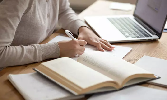 student writing in composition book with laptop nearby