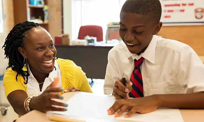 A smiling teacher kneels beside a student to discuss his work, representing the difference between teaching online vs in a classroom