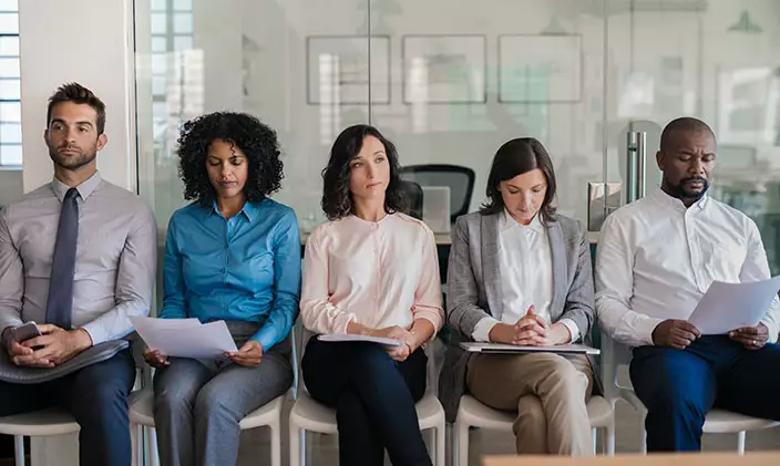 A group of job applicants await interviews, resumes in their hands.
