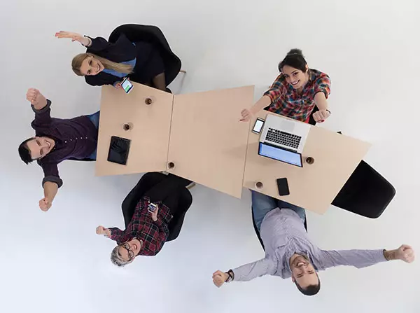 Colleagues seated at a table raise their hands and look to the ceiling enthusiastically