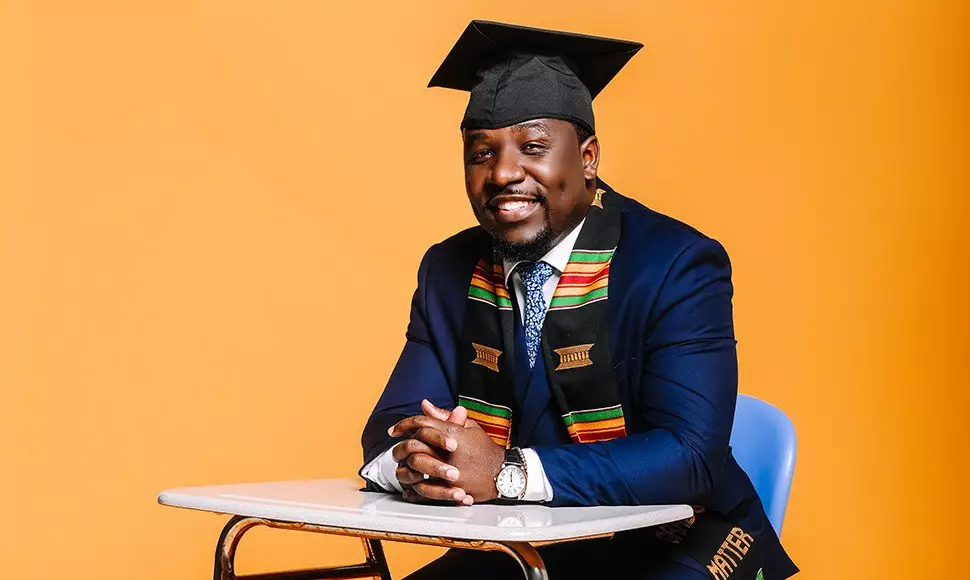 Jahmar Robinson sits in a high school class desk wearing his graduation regalia