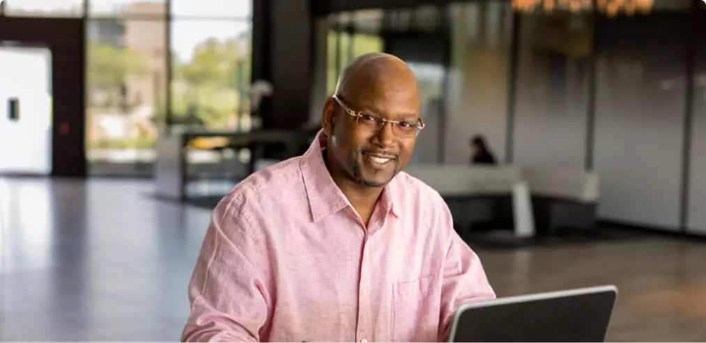 A person in a pink shirt smiling while using a laptop indoors, suggesting an online learning environment.