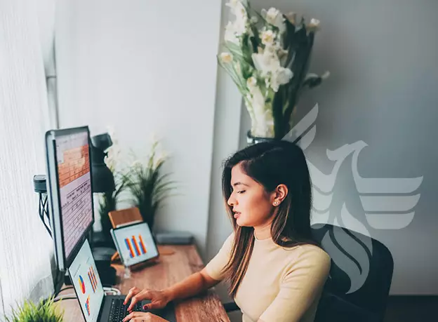 A business woman sitting at a desk reviewing three monitors of graphs and information