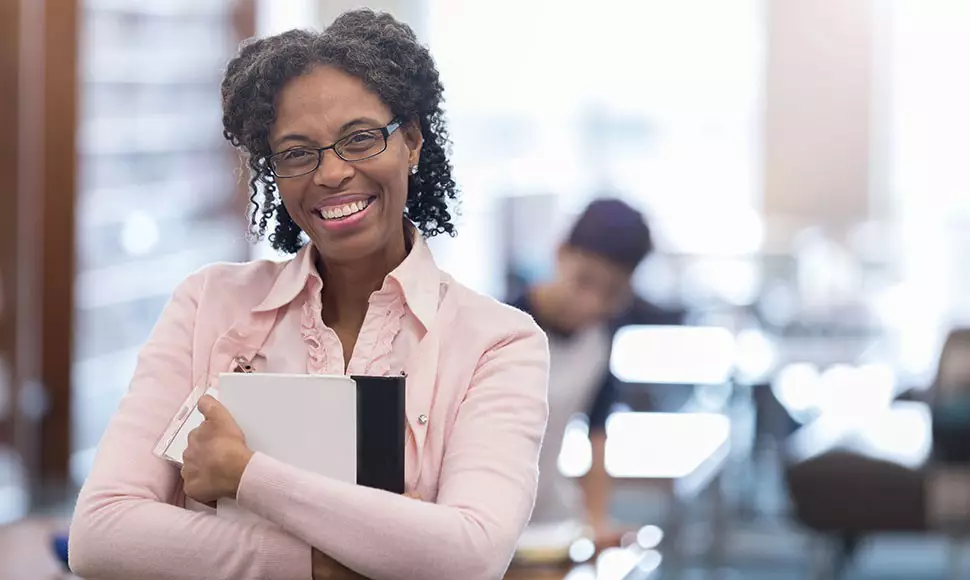 Older female worker in a pink collared shirt smiles in a window-walled office and represents the topic ageism