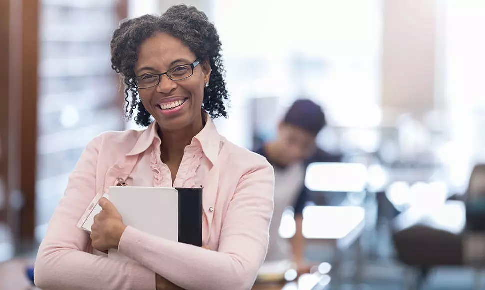 Mature Black woman clutches a notebook while smiling in an office