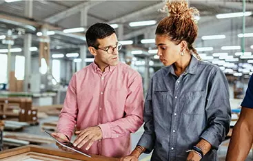 An operation manager and colleague stand at a table in an open office area, checking a tablet