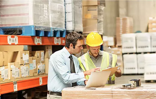 An operations manager in a warehouse meets with an employee wearing a hard hat