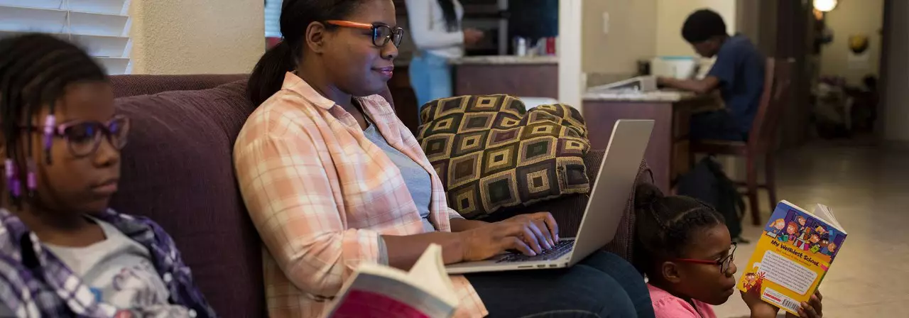 A mother works on a laptop from her couch with three children sitting nearby also working and reading.
