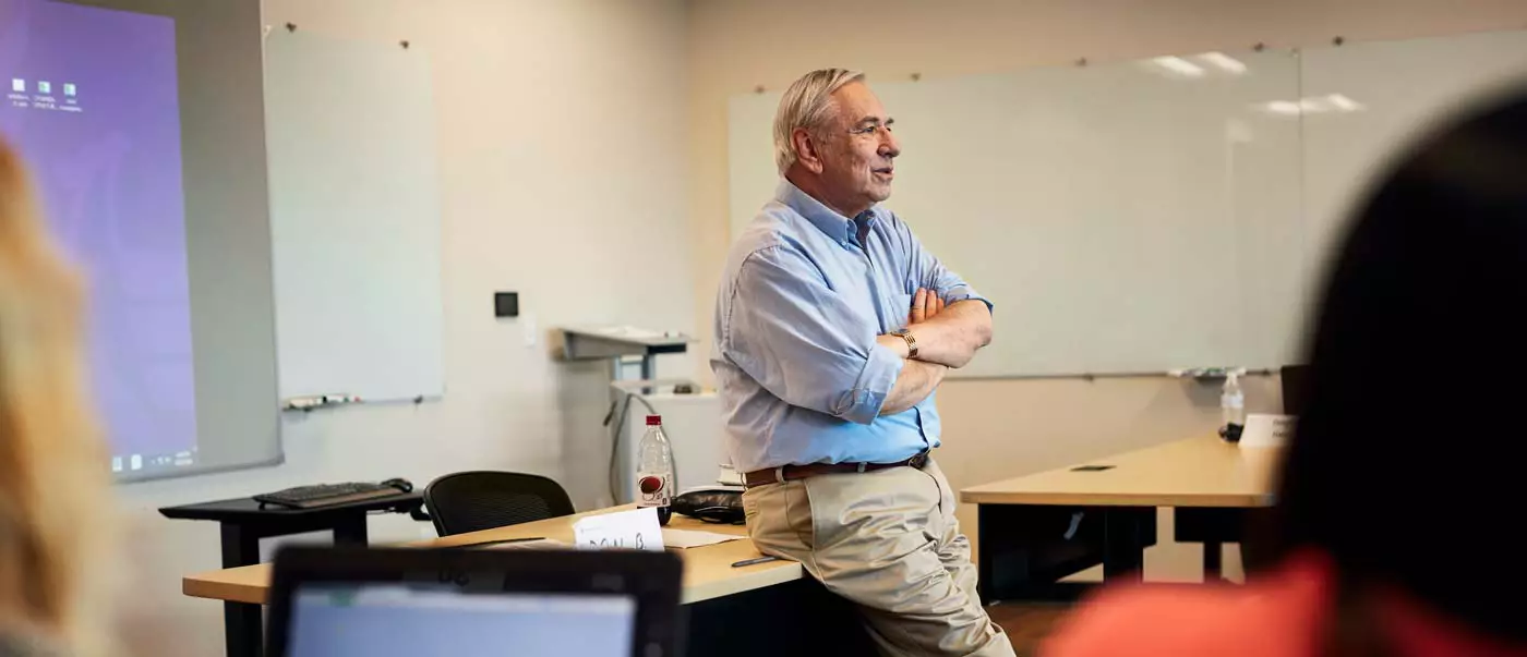 A lecturer leans on a desk and chats with his students