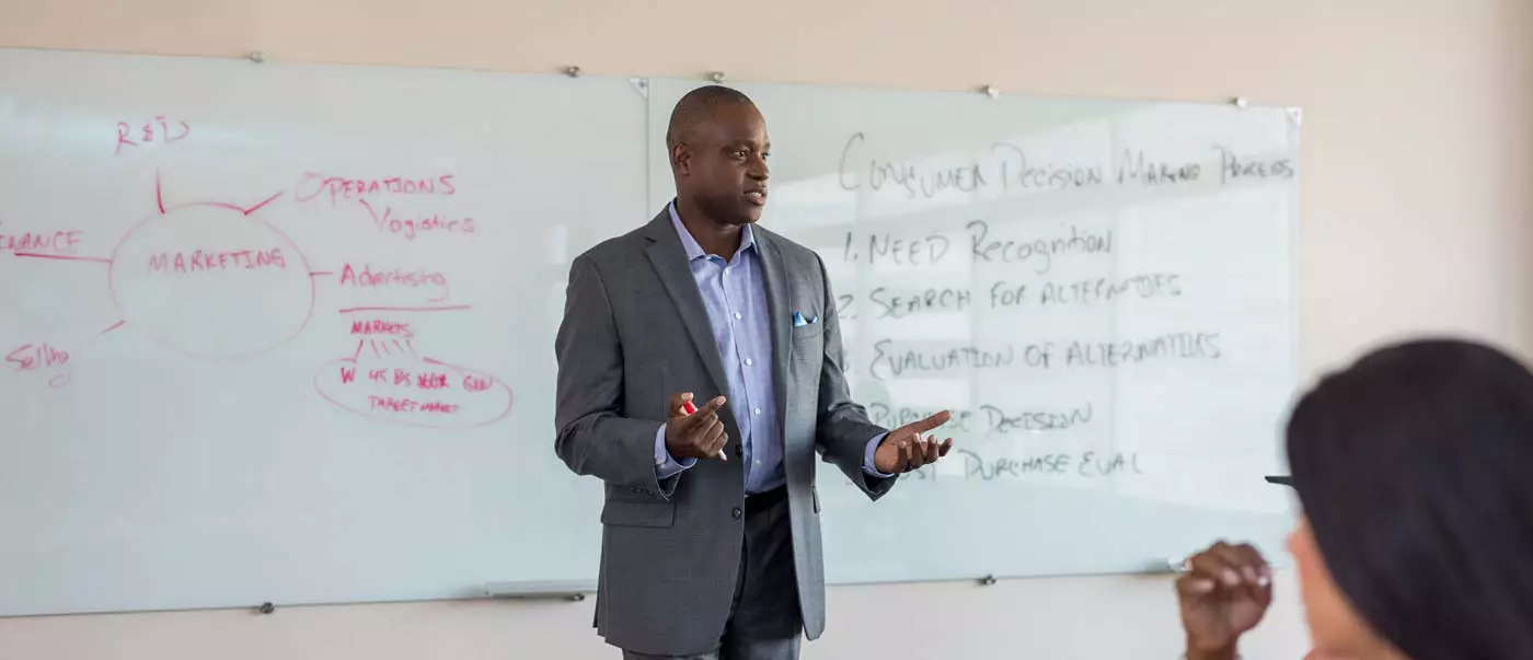 A lecturer speaks to a class while standing in front of a whiteboard