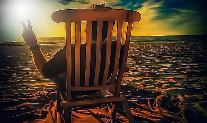 A photo of the beach and the back of a man laid back in a chair holding up a piece sign against the scenery
