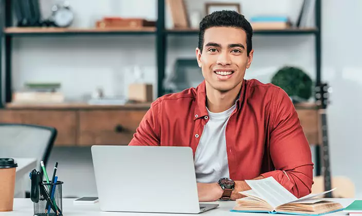Male student taking online classes on laptop with open book next to him