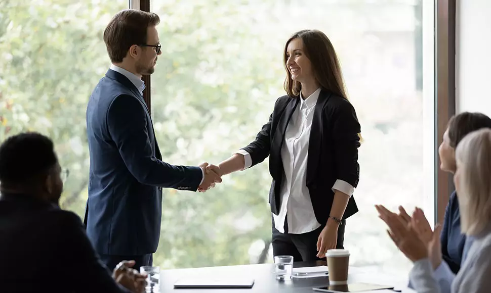 Man and woman shake hands in front of a conference table of clapping people